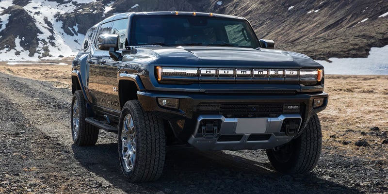 Dark gray GMC Hummer EV SUV parked on a gravel road in a rugged landscape with snow-capped mountains.