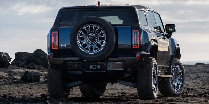 Rear view of a dark blue Hummer EV parked on dark, rocky ground, with a large spare tire on its back tailgate.