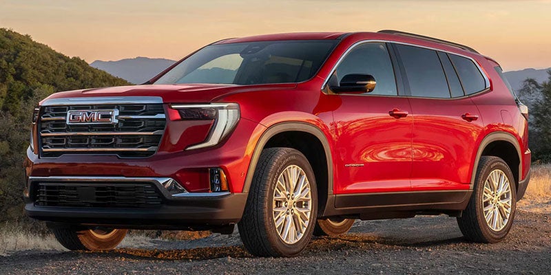 A red GMC Acadia AT4 SUV parked on a dirt road with mountains and sunset visible in the background.