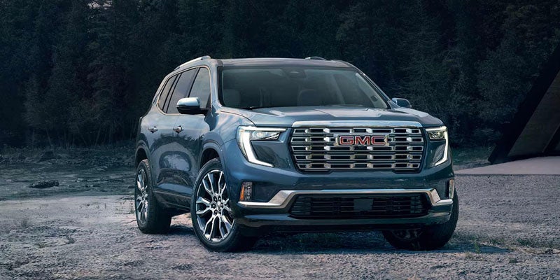 A dark blue GMC Acadia SUV, with prominent headlights and chrome grille, parked on rough terrain with trees in the background.