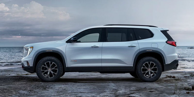 A light grey GMC SUV with black trim and black wheels parked on a rocky beach by the sea under a cloudy sky.