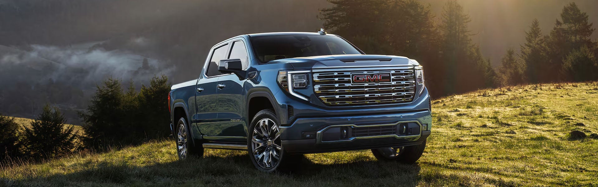 A blue GMC Sierra pickup truck parked on a dewy, grassy hill with pine trees and misty mountains in the background.