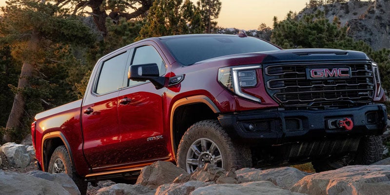 A red GMC Sierra AT4X pickup truck angled on a rocky trail with trees and a mountain in the background.