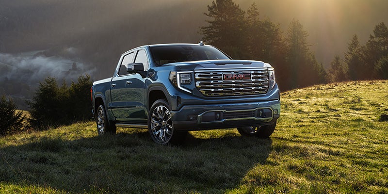 Blue GMC Sierra AT4X truck parked on a grassy hill with trees and misty mountains in the background.