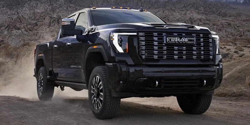 A black GMC Denali HD pickup truck driving on a dusty dirt road with rocky mountains in the background.