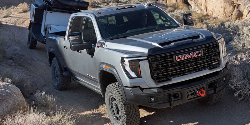 A silver GMC Sierra pickup truck towing a utility trailer on a rocky, desert dirt road.