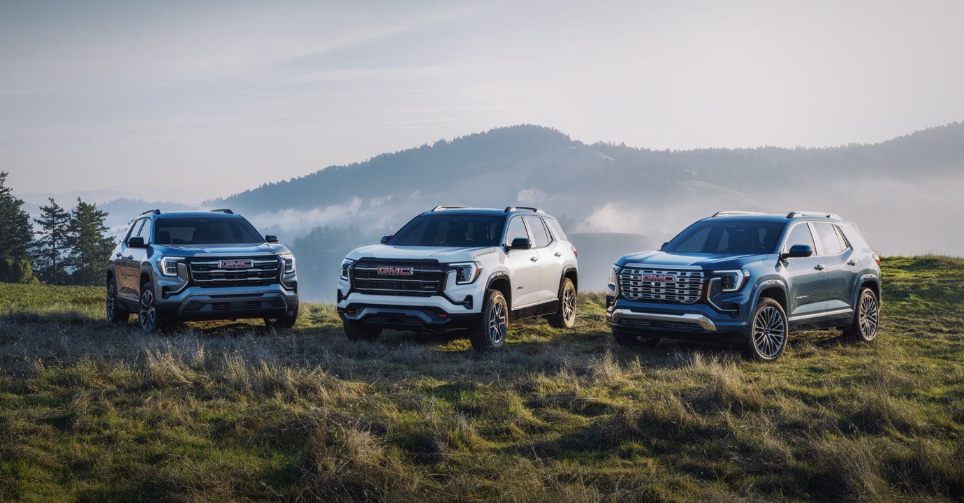 Three GMC Terrain SUVs, dark gray, white, and blue, parked left to right on a grassy hill with mountains in the background.