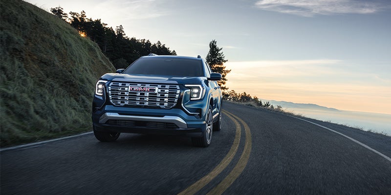 Front view of a dark blue GMC SUV with bright headlights on a winding road, with a sunset sky and mountains.