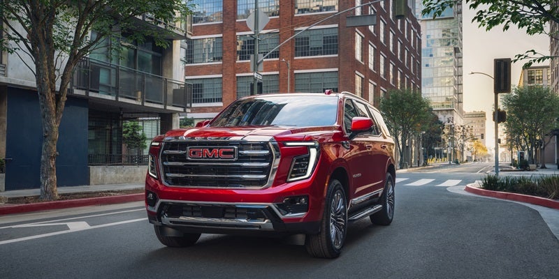 A glossy red GMC Yukon SUV parked on a city street, with urban buildings and trees in the background.