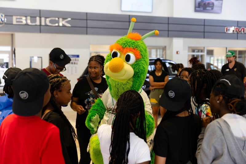 Green caterpillar mascot in a baseball jersey surrounded by children in a car dealership.